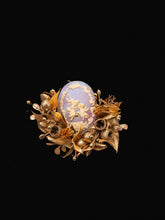 The Melaleuca Bracteata Birth Flower Ring by CAMELLIA is an ornate decorative piece with floral and gold embellishments, handcrafted in New York. It features a striking combination of white birds amidst clouds on a blue background set against a black backdrop.
